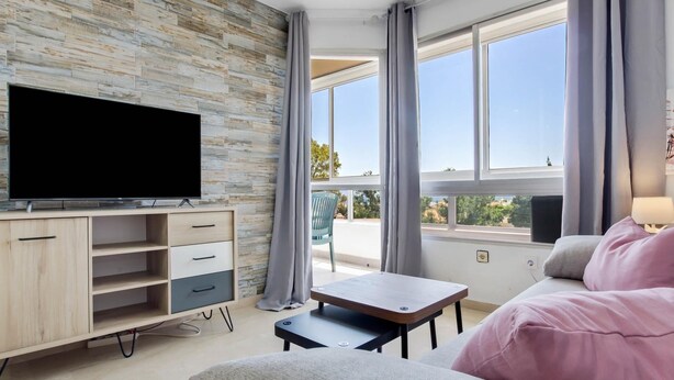 Una sala de estar moderna con un televisor de pantalla plana, estanterías de madera, una mesita, un sofá y ventanas amplias que ofrecen vistas al exterior.