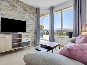 Una sala de estar moderna con un televisor de pantalla plana, estanterías de madera, una mesita, un sofá y ventanas amplias que ofrecen vistas al exterior.