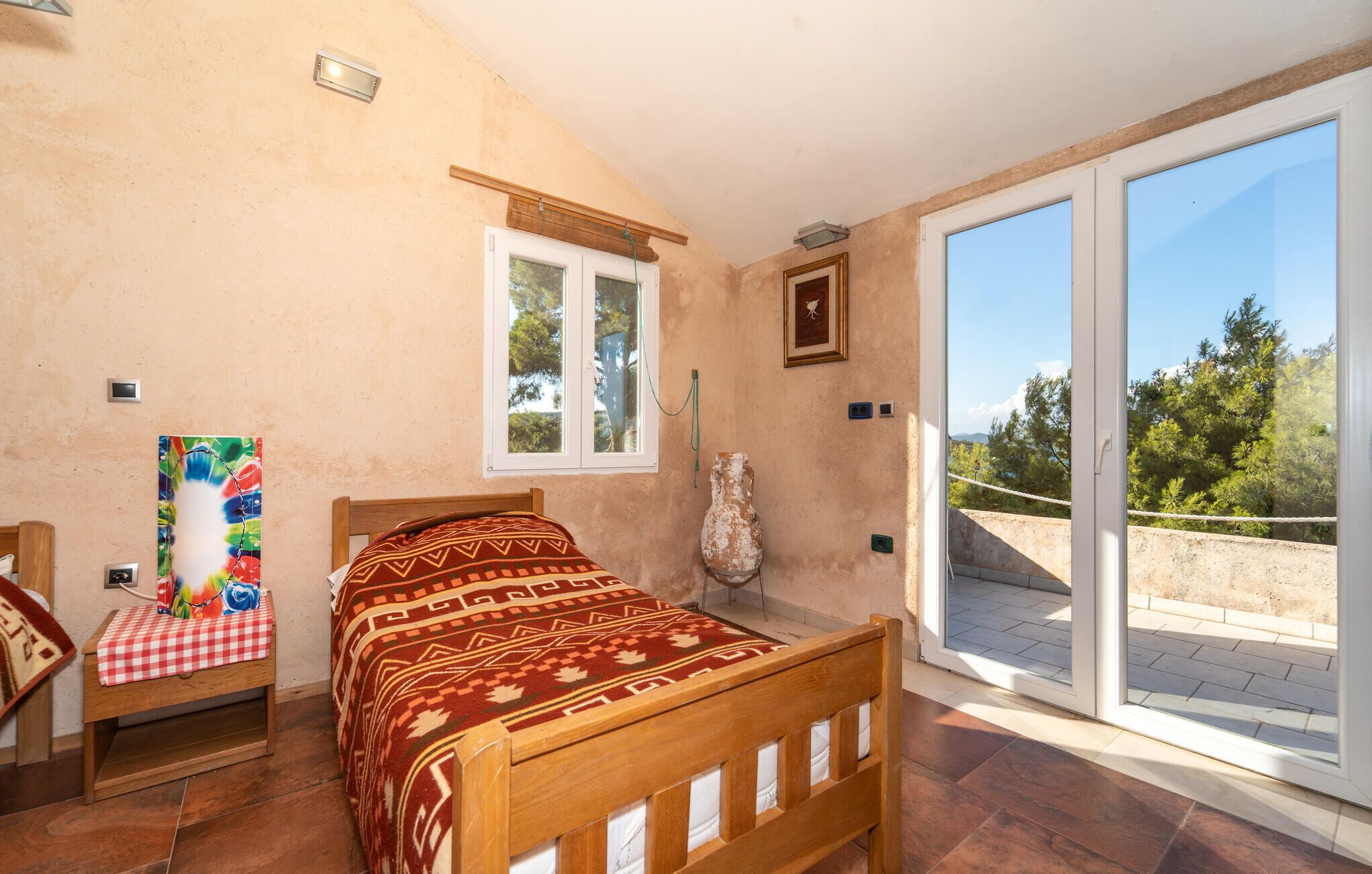 Ein Schlafzimmer mit einem hölzernen Bett, einem karierten Tisch, einem farbenfrohen Gemälde und Blick auf einen Balkon mit Bäumen.
