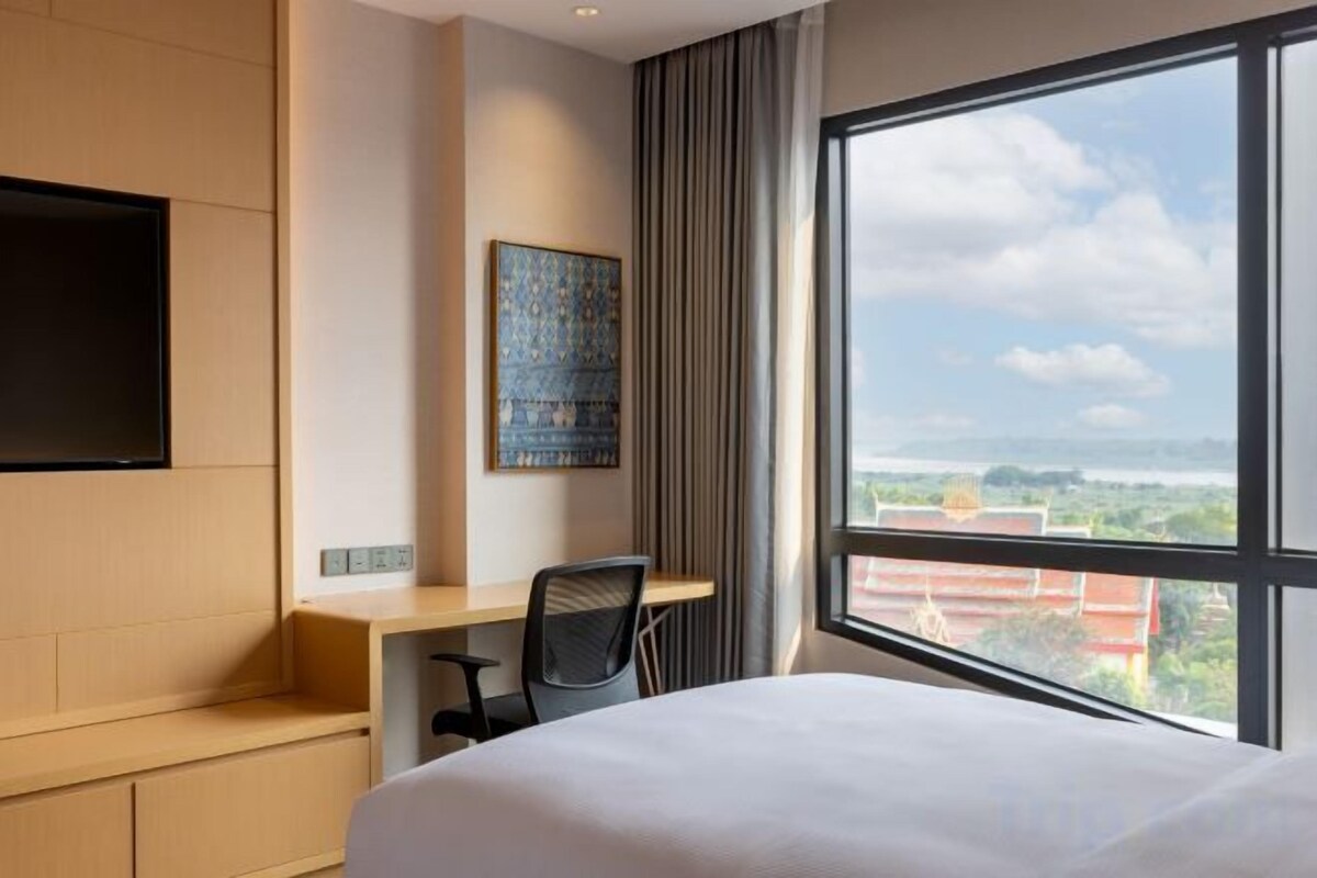 A hotel room with a large bed, a desk, a chair, a flat-screen TV mounted on the wall, and a large window offering a city view.