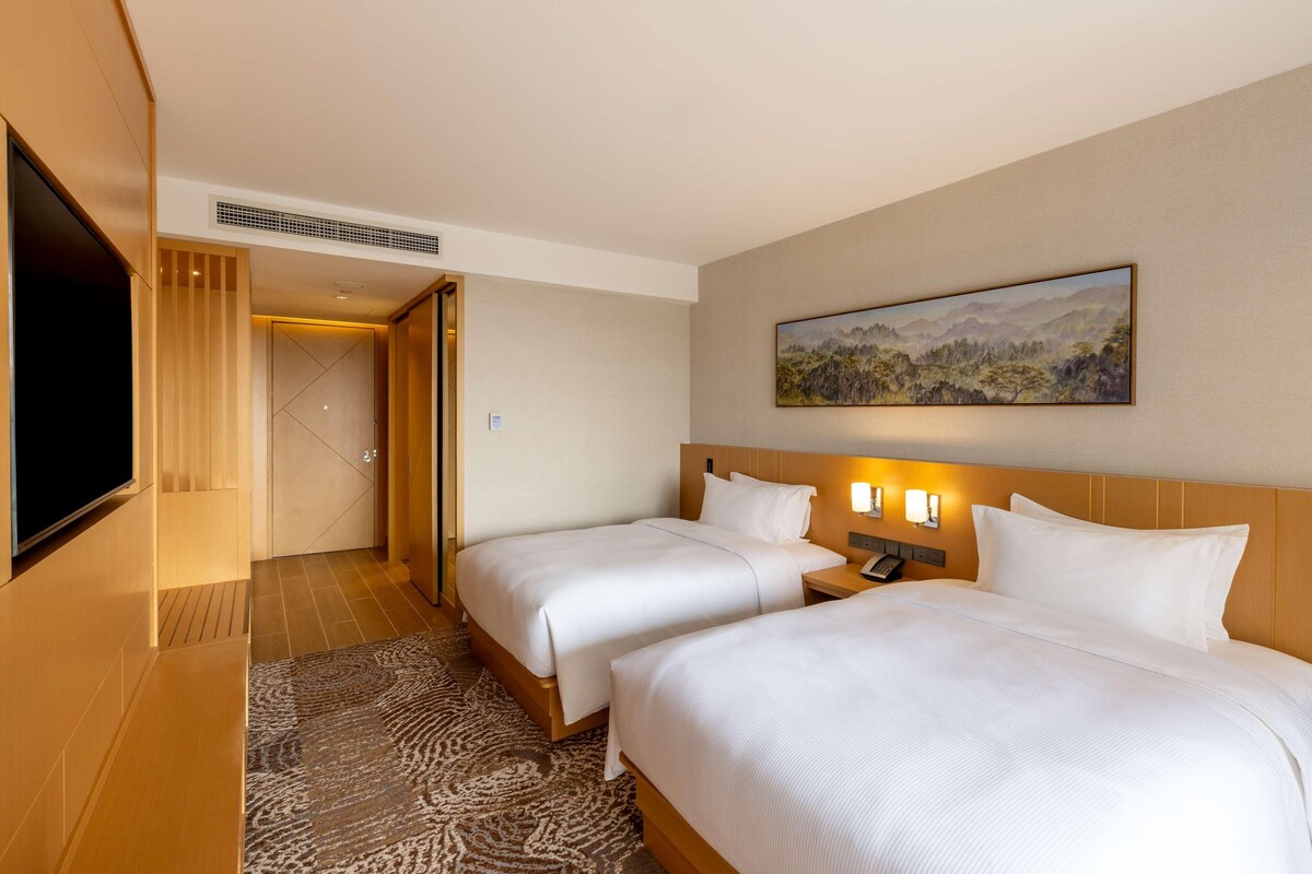 A hotel room with two beds, a flat-screen TV, a painting on the wall, and a wooden headboard.