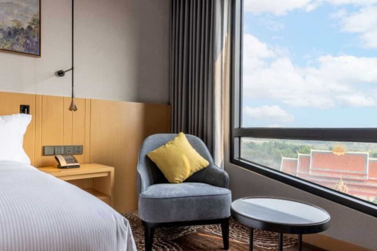 A hotel room with a bed, a chair with a pillow, a small table, a nightstand with a phone, and a window with a view of buildings.