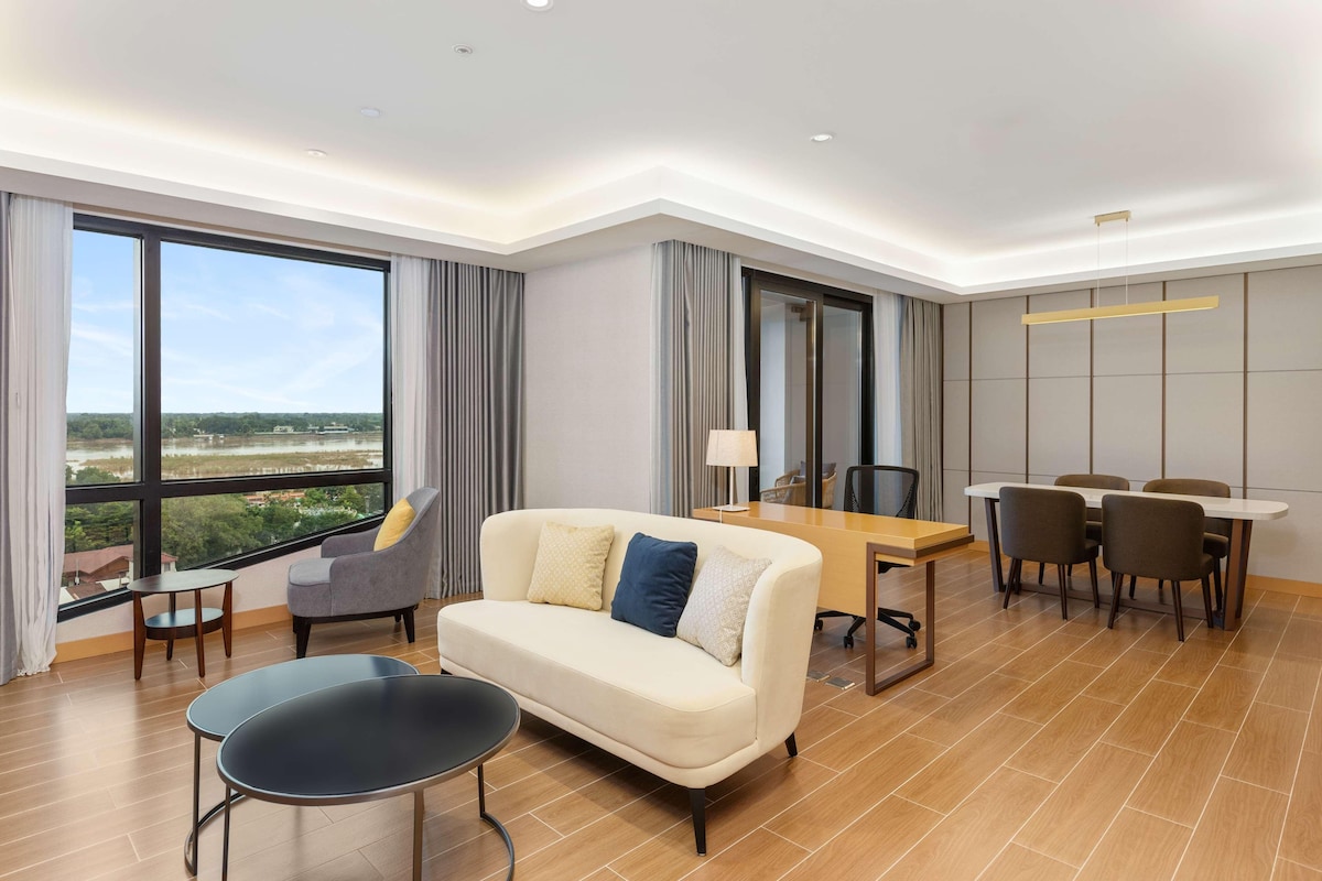 Een moderne hotelkamer met een groot raam, een bank, een eettafel met stoelen en een klein bijzettafeltje.