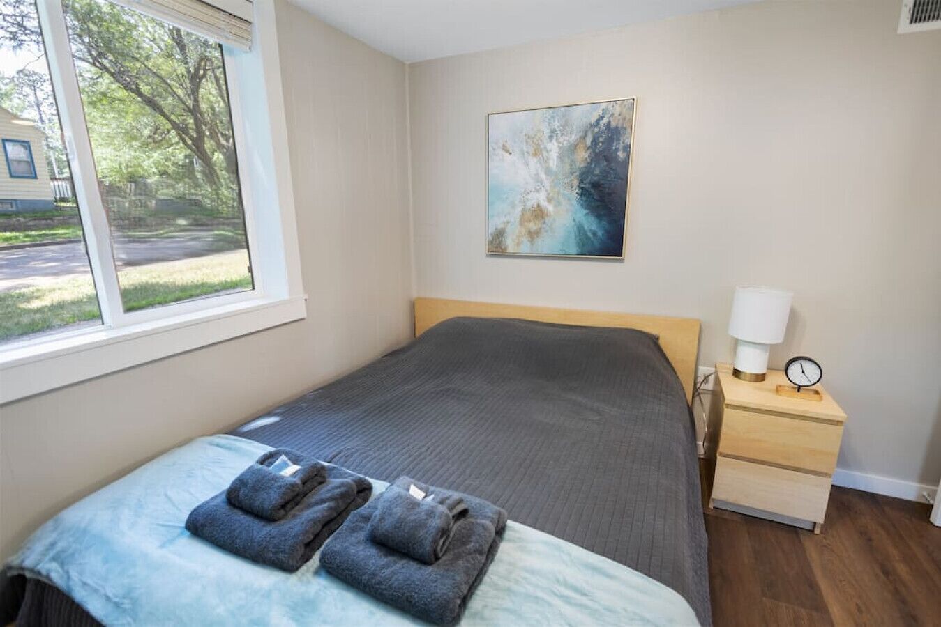 A bedroom with a bed, a nightstand with a lamp, a painting on the wall, and a window with a view of a house and trees.
