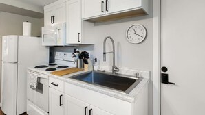 A compact kitchen with white cabinets, a stainless steel sink, a microwave, a stove, and a clock on the wall.