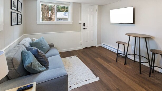 A living room with a grey sofa, a white rug, a TV mounted on the wall, and a door leading outside.