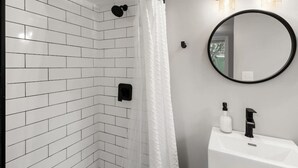 A modern bathroom with white subway tiles, a shower with a white curtain, a round mirror, and a white sink with black fixtures.