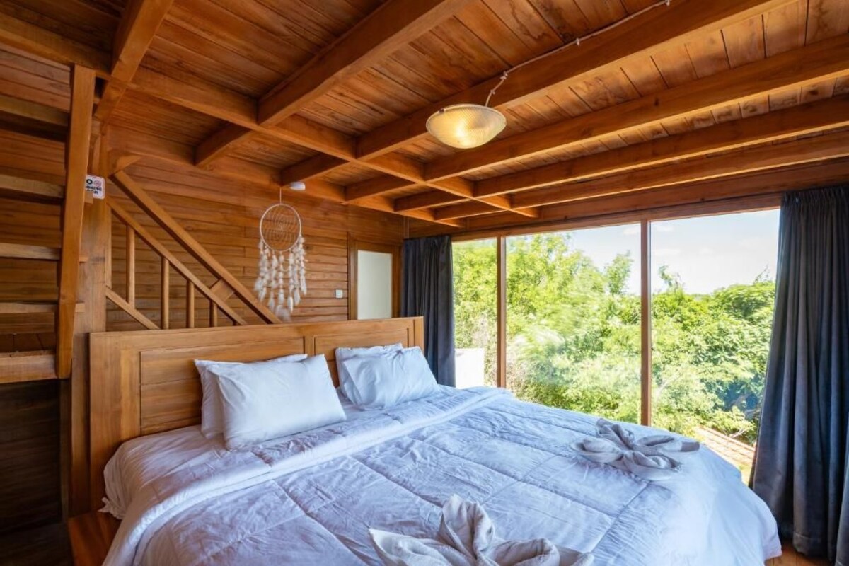 A wooden cabin bedroom with a large bed, a hanging dreamcatcher, and a view of greenery outside.