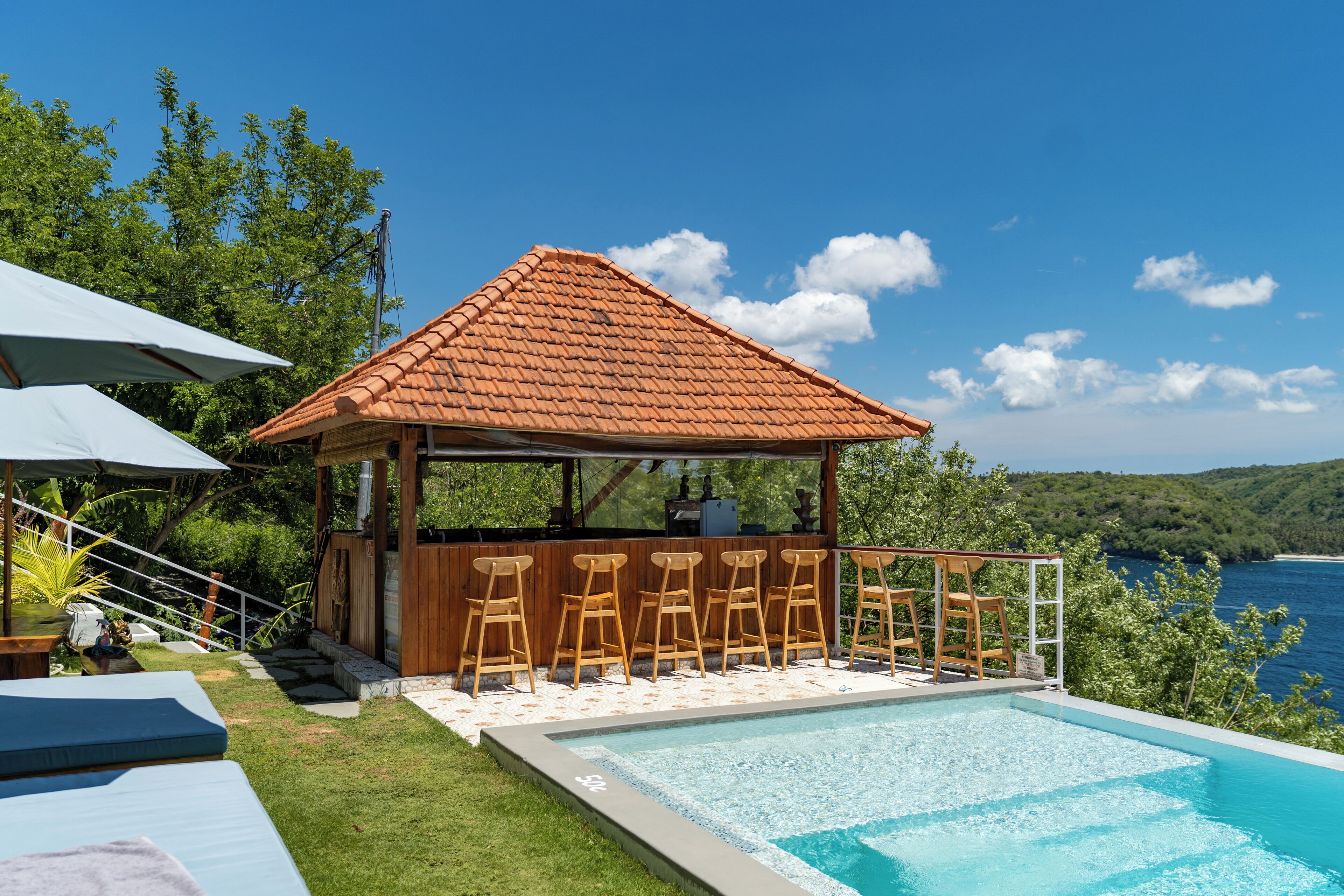 Uma área à beira da piscina com um gazebo de madeira, espreguiçadeiras e vista para o mar.