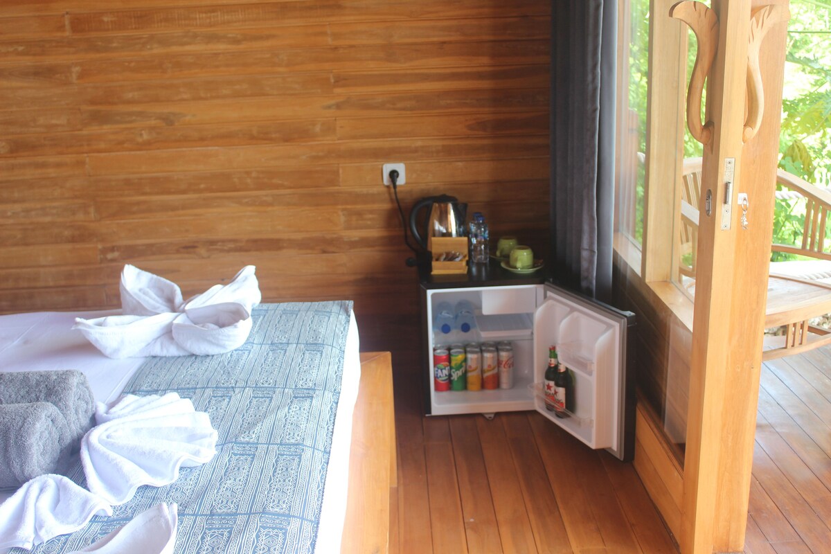 A wooden cabin room with a bed, a small refrigerator stocked with beverages, and a view of the outdoors.