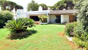 Una propiedad con fachada blanca, techo de tejas y un jardín cuidado con diversas plantas y una palmera.