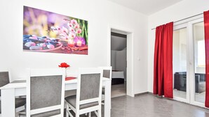 A dining area with a white table and chairs, a red flower centerpiece, and a painting of flowers and rocks on the wall. - Apartment Zubčić by Interhome (Sv. Filip i Jakov)