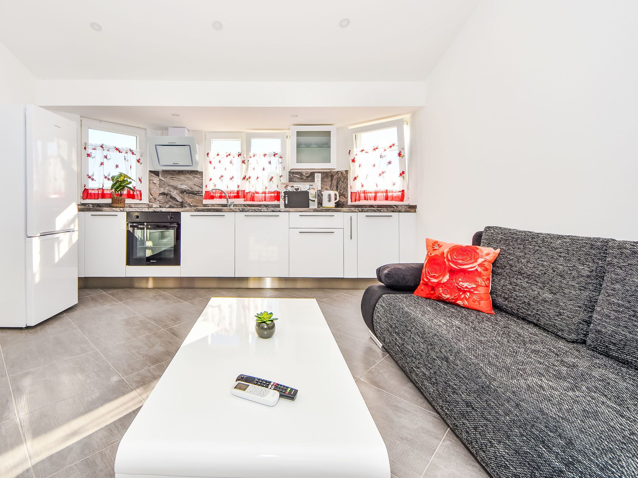A modern kitchen with white cabinets, a built-in oven, and a microwave. There is a white coffee table with a remote control and a small plant.