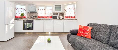 A modern kitchen with white cabinets, a built-in oven, and a microwave. There is a white coffee table with a remote control and a small plant.