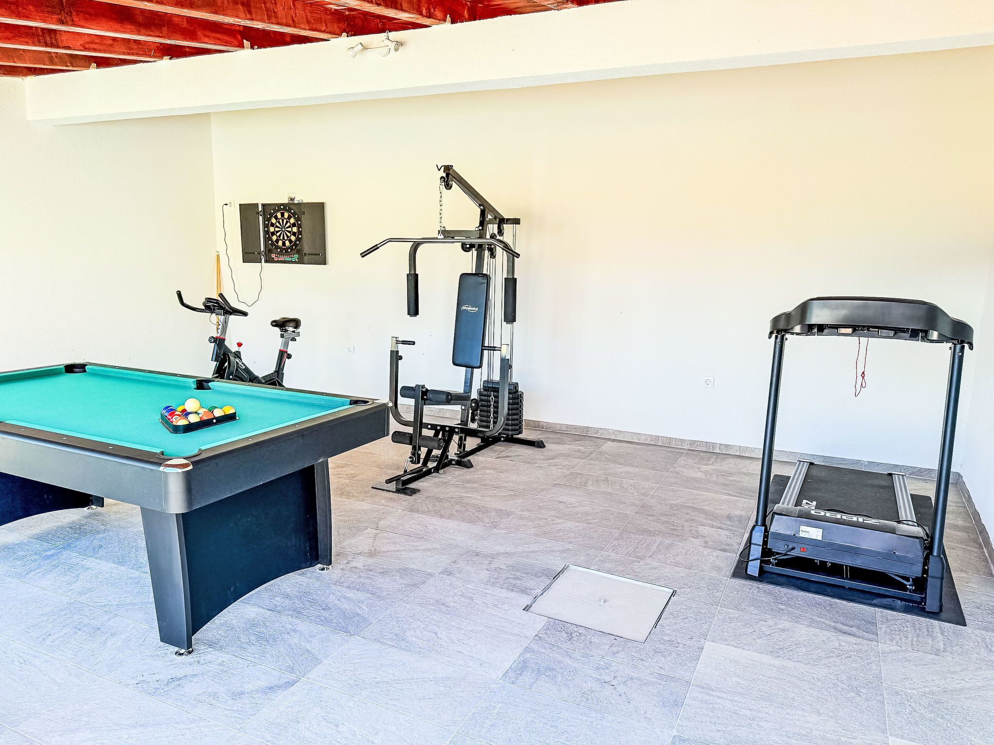 Una mesa de billar con bolas, una cinta de correr y equipo de ejercicio en una habitación con piso de baldosas y paredes blancas.