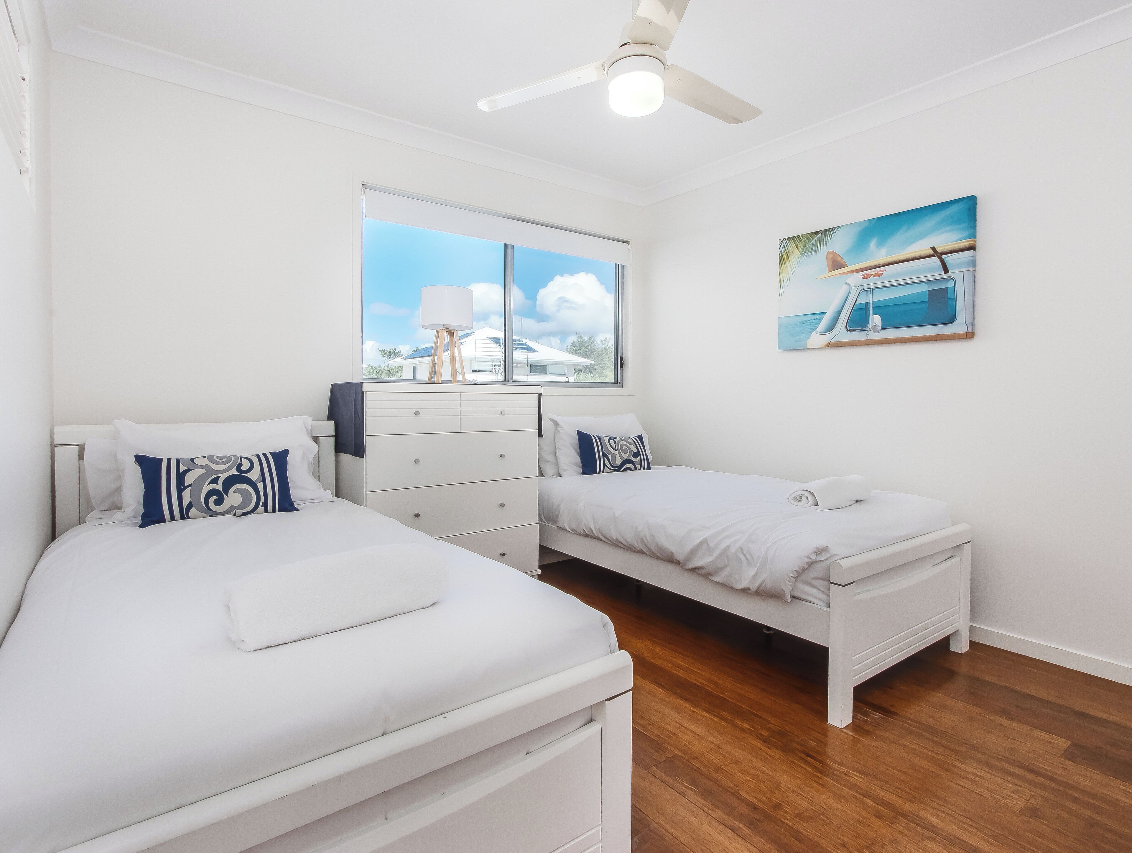 A bedroom with two beds, a ceiling fan, a window with a view, and a painting on the wall.