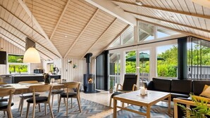 Un vaste espace de vie doté d’un plafond en bois, d’une grande fenêtre, d’une cheminée et d’un coin repas avec une table et des chaises.