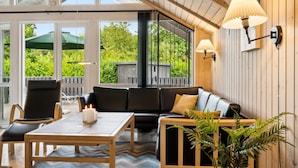 Un salon chaleureux avec un plafond en bois, un canapé en cuir noir, une table basse en bois et une grande fenêtre donnant sur une terrasse en bois et un espace verdoyant.