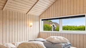 Une chambre avec un plafond et des murs en bois, un lit avec une literie blanche et bleue, et une fenêtre donnant sur la verdure.