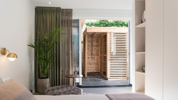 Une chambre moderne avec une grande fenêtre, une plante en pot, une table de chevet avec des lunettes, et une porte en bois donnant sur un espace douche.