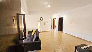 Uma sala de estar ampla com um sofá, uma mesinha e um piano. Existem duas portas que dão para outros cômodos.