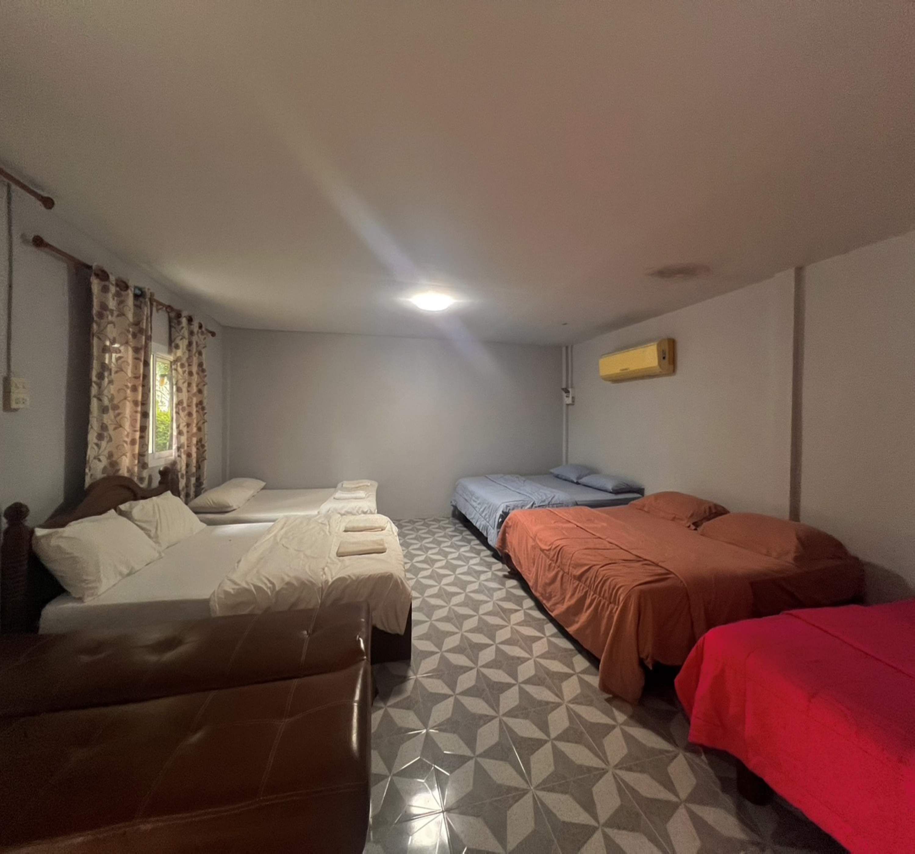 A room with four beds, a patterned floor, a window with curtains, and a ceiling light.