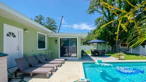 A pool with lounge chairs, a covered patio, and a house with a sliding glass door.