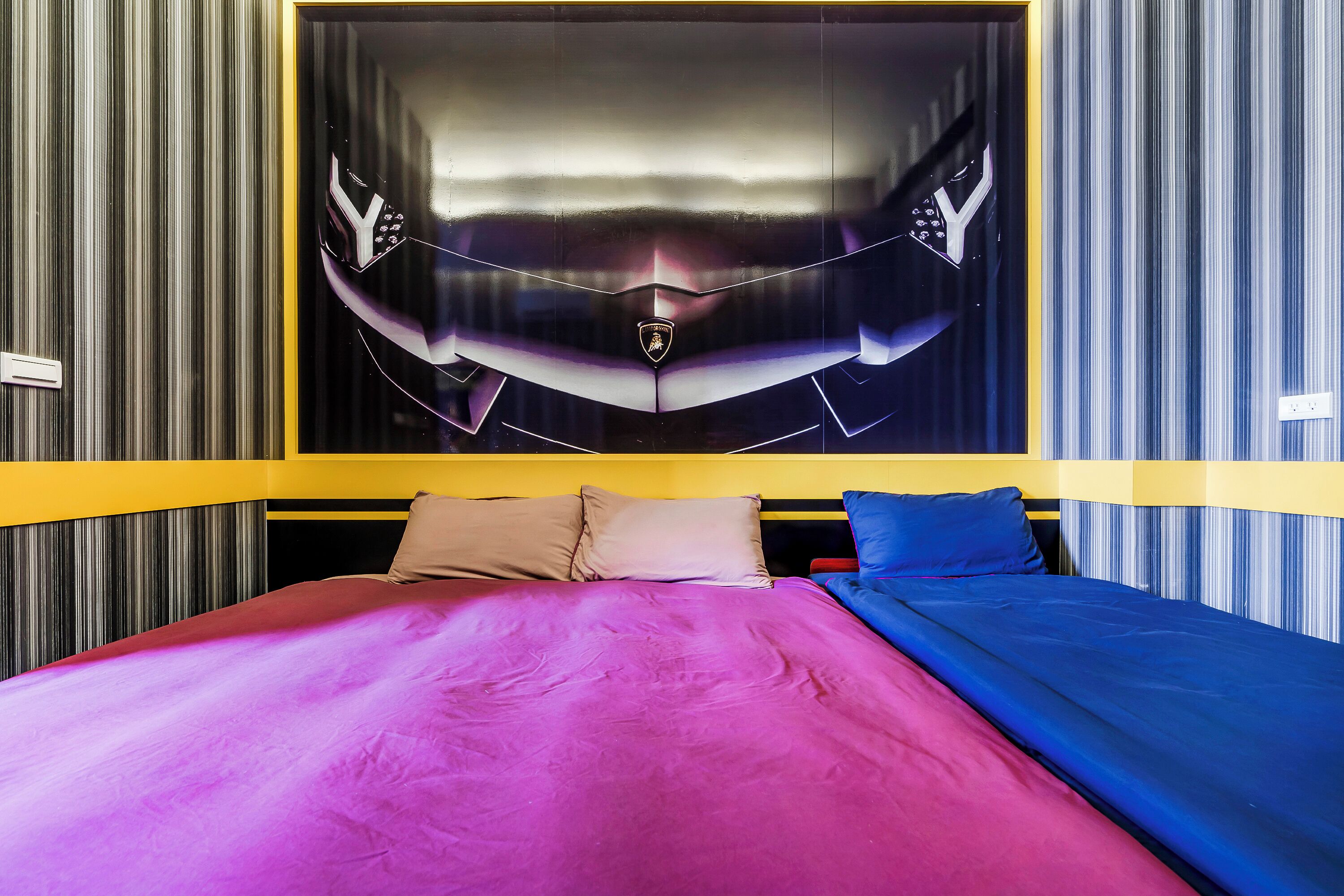 A bed with a pink and blue bedspread, a headboard with a yellow accent, and a wall with vertical stripes.