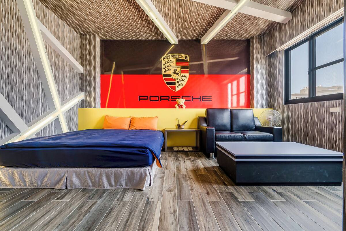Ein modernes Zimmer mit einem Bett, einem Schlafsofa und einem kleinen Tisch. An der Wand ist ein großes Porsche-Logo zu sehen.