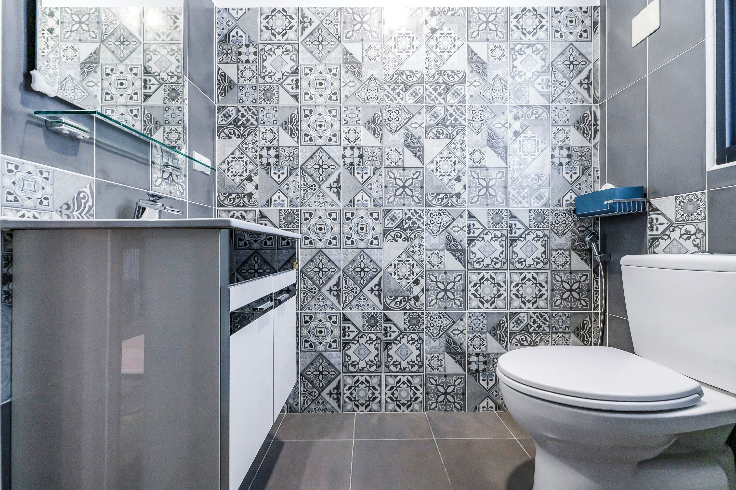 A bathroom with a white toilet, a glass shelf, and a tiled wall with geometric patterns.