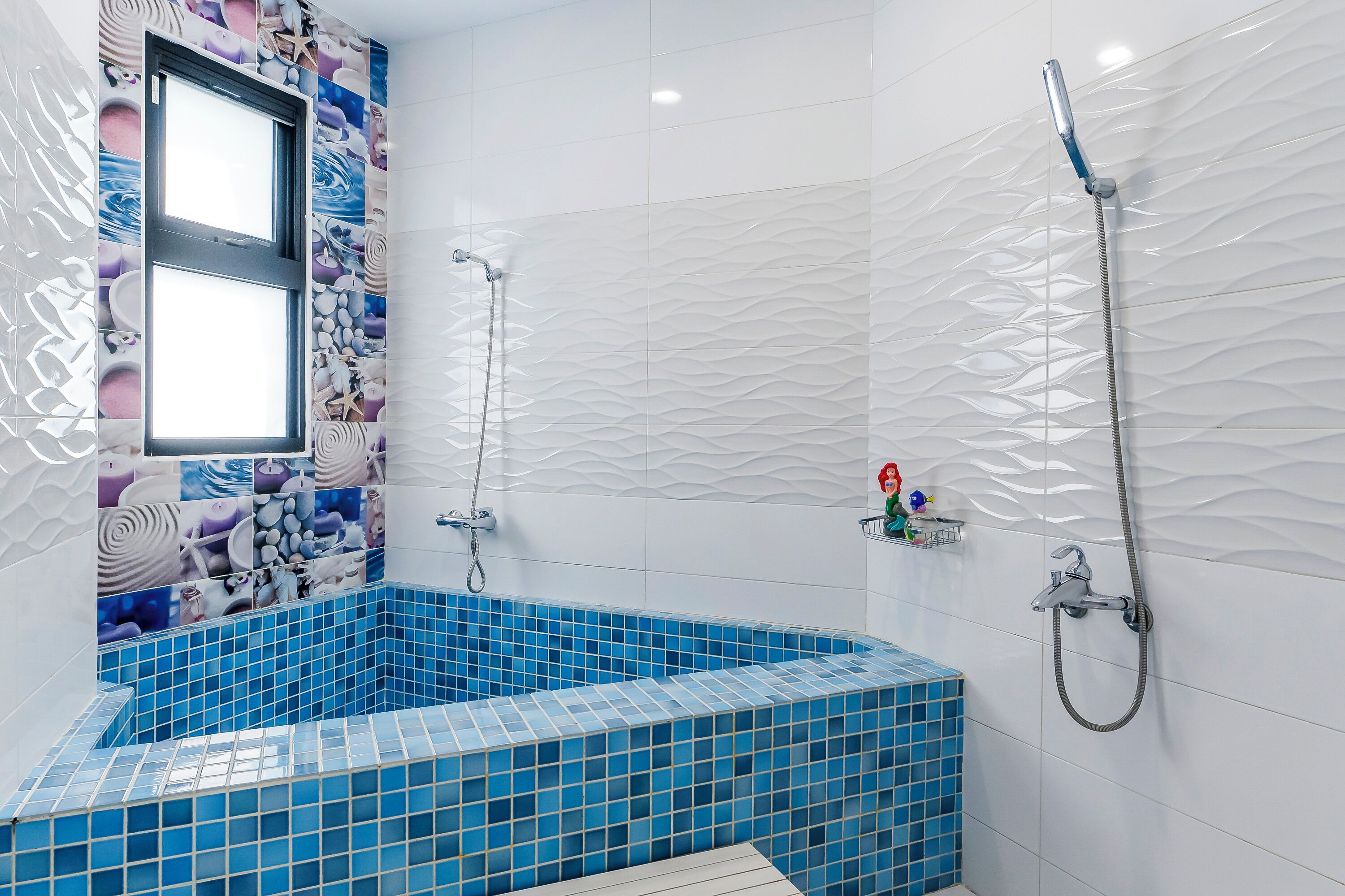 A bathroom with a blue-tiled bathtub, a shower with a handheld showerhead, and a window with a view of the sea.