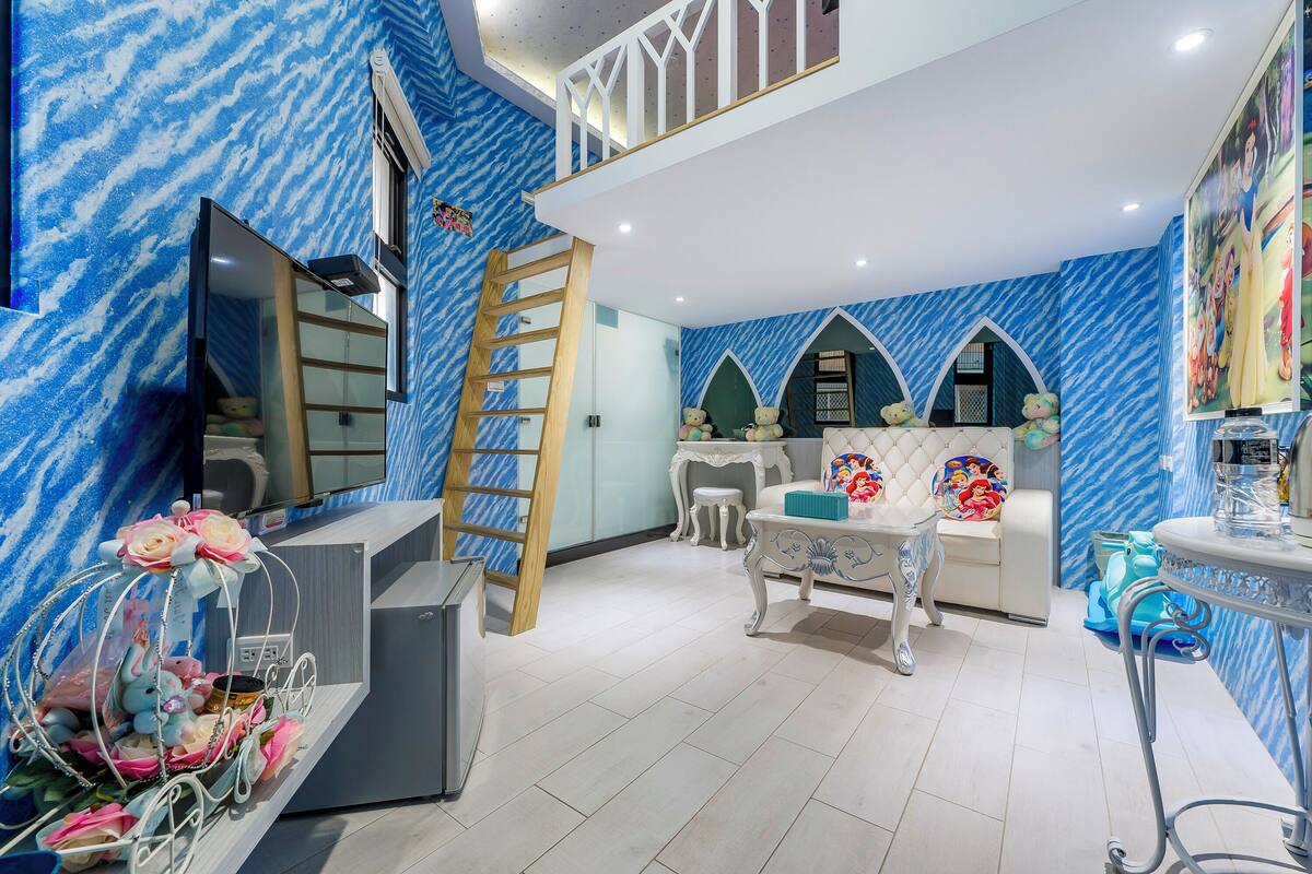 Ein Zimmer mit blauer Wandgestaltung, einem Schlafbereich mit Leiter, einem weißen Sofa, einem kleinen Tisch und einem dekorativen Regal mit Stofftieren.