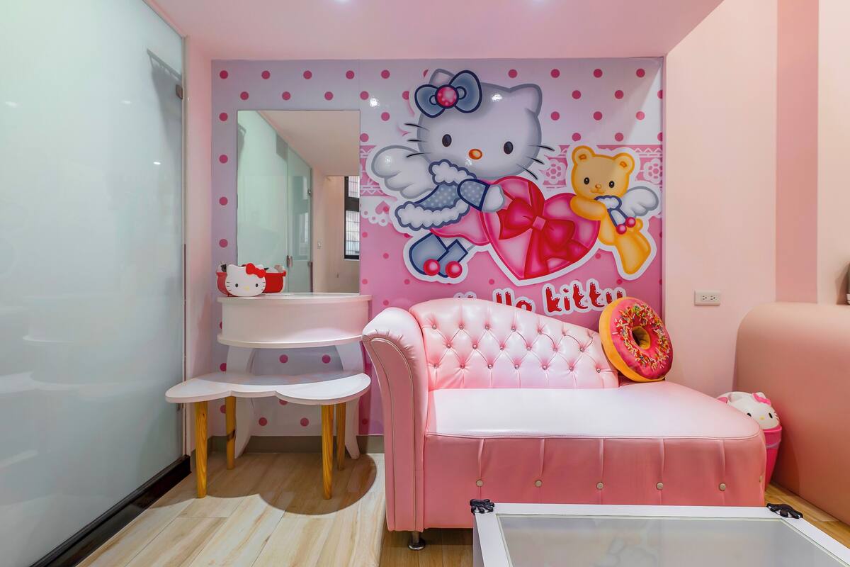 Ein zartrosa Zimmer mit einer Hello Kitty Wandmalerei, einem rosafarbenen Sofa, einem kleinen weißen Tisch und einem rosa Sessel.