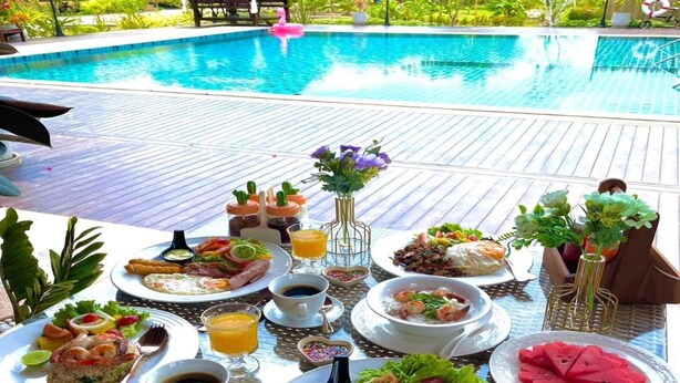 A dining table with various dishes and drinks by a poolside, overlooking a lush garden and a pool. - Capital O 75518 Baan Rose Resort Ao Luk (Ao Luek)