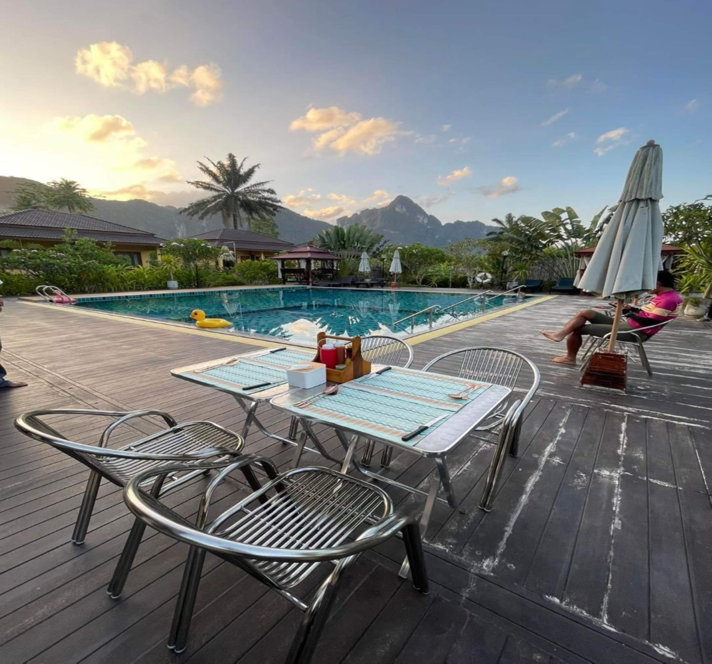 A poolside area with a wooden deck, metal chairs, a table with a menu, and a view of mountains in the background.