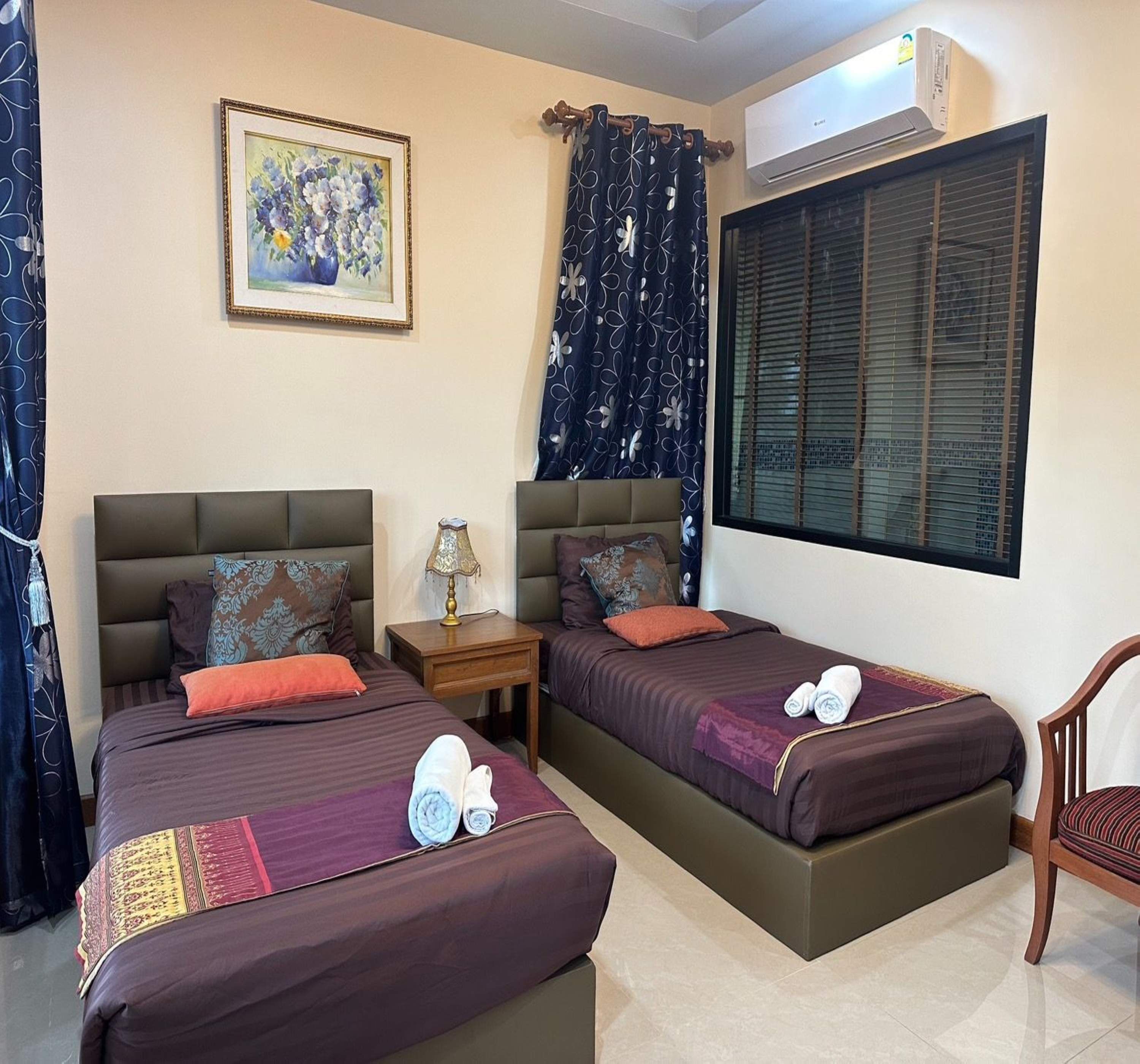A hotel room with two beds, a nightstand, a lamp, a painting, and an air conditioning unit.