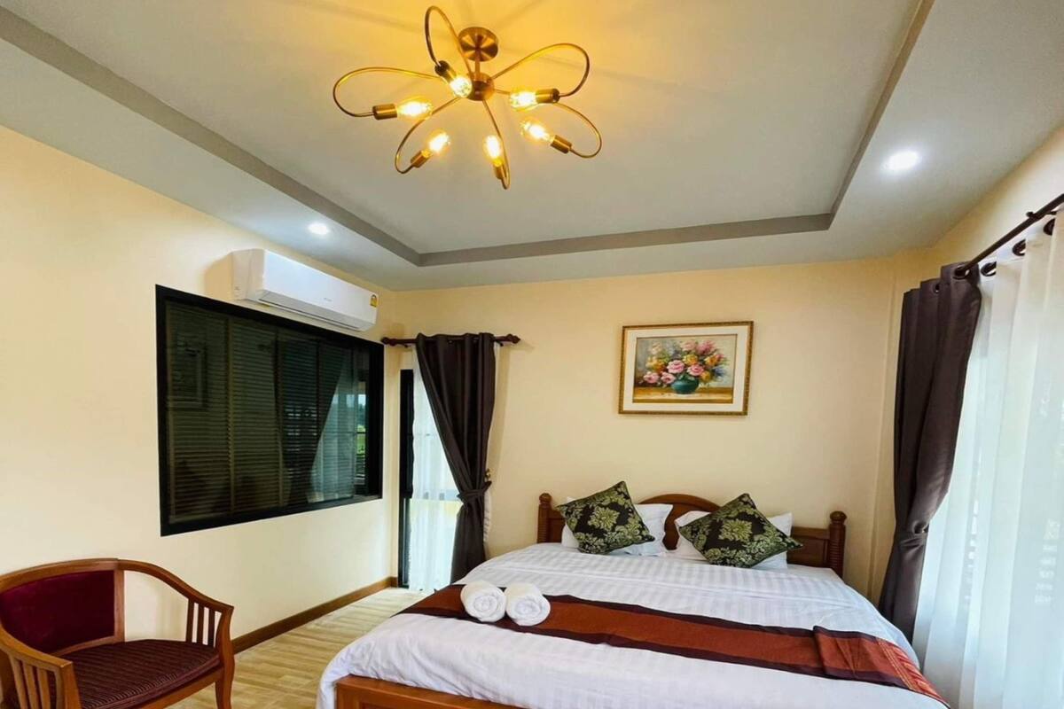A neatly arranged bedroom with a wooden bed, a chair, a ceiling light fixture, and a window with curtains.