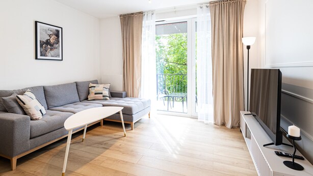 Un salon moderne avec un canapé gris, une table basse blanche, un téléviseur à écran plat et un balcon agrémenté de rideaux.