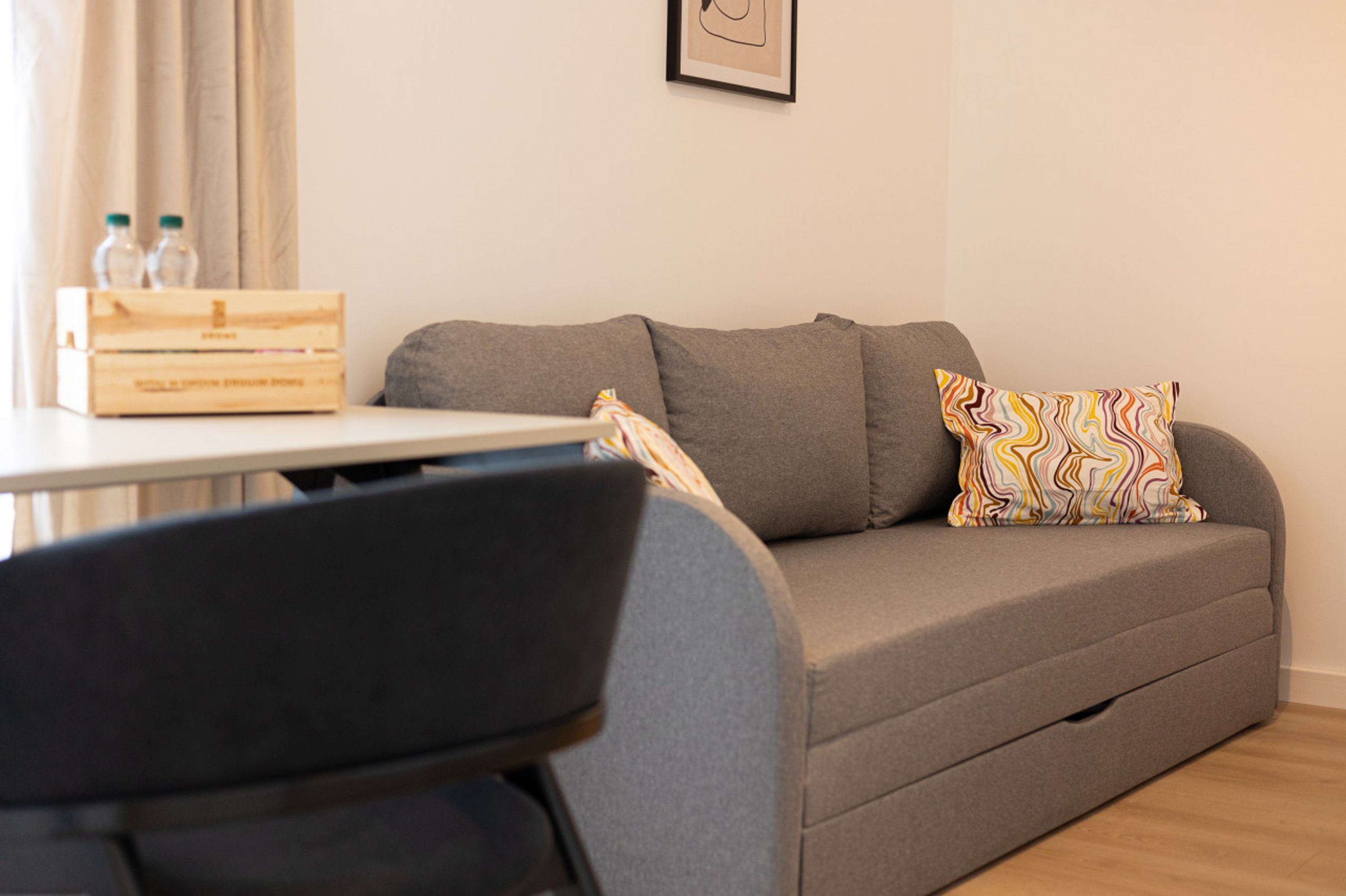 Ein graues Sofa mit einem farbenfrohen Kissen, eine Holzkiste mit Flaschen und ein gerahmtes Bild an der Wand.
