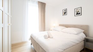 Un lit bien fait, avec du linge de lit blanc et une serviette pliée, une table de chevet avec une lampe, et deux tableaux encadrés accrochés au mur.