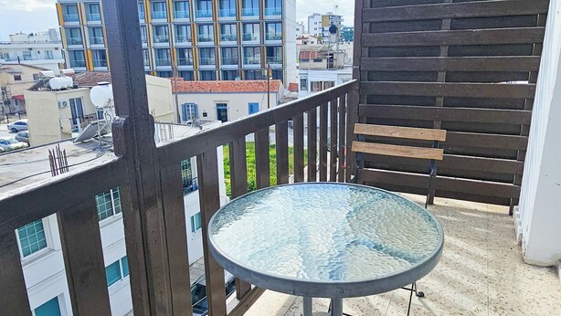 A balcony with a glass-top table and wooden benches, overlooking a cityscape with buildings and a clear sky. - Downtown Rooms, 200m from Marina Beach (Larnaca)