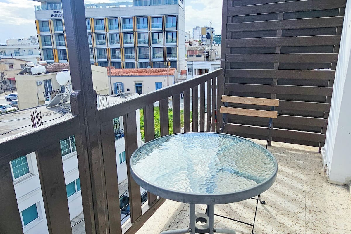 Uma varanda com mesa de vidro e bancos de madeira, com vista para um horizonte urbano com edifícios e céu limpo.