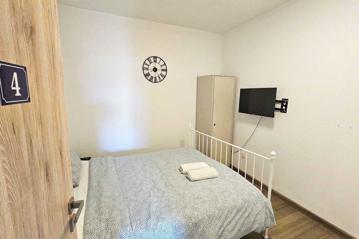 Quarto pequeno e simples com uma cama, um relógio, uma televisão e uma porta com o símbolo de número.