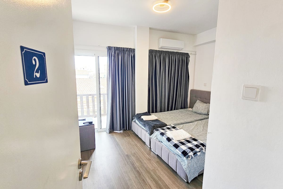 Quarto de hotel com cama, uma mesinha e uma cadeira. Há uma janela com cortinas e um aparelho de ar condicionado na parede.