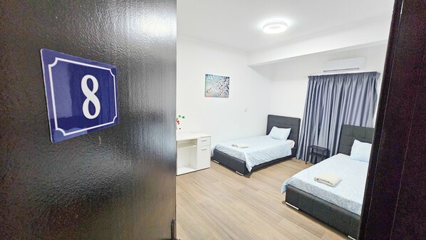 A hotel room with two beds, a desk, a chair, a painting on the wall, and a number sign on the door. - Downtown Rooms, 200m from Marina Beach (Larnaca)