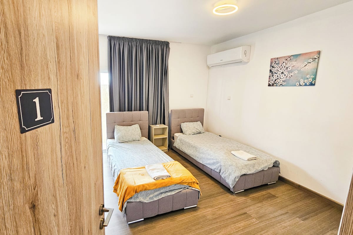 Quarto com duas camas, porta de madeira com um número, ar condicionado e uma pintura na parede.