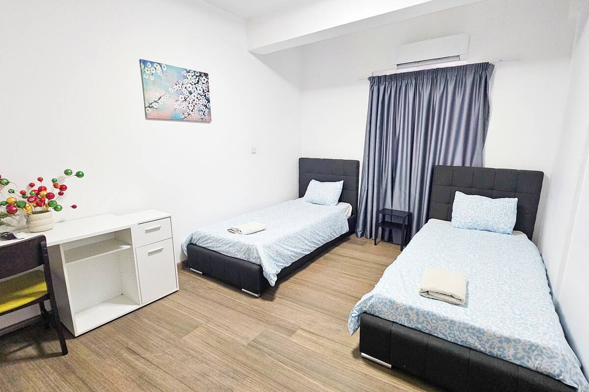Quarto de hotel com duas camas de solteiro, uma escrivaninha com cadeira, uma mesinha, um vaso de planta e um quadro na parede.