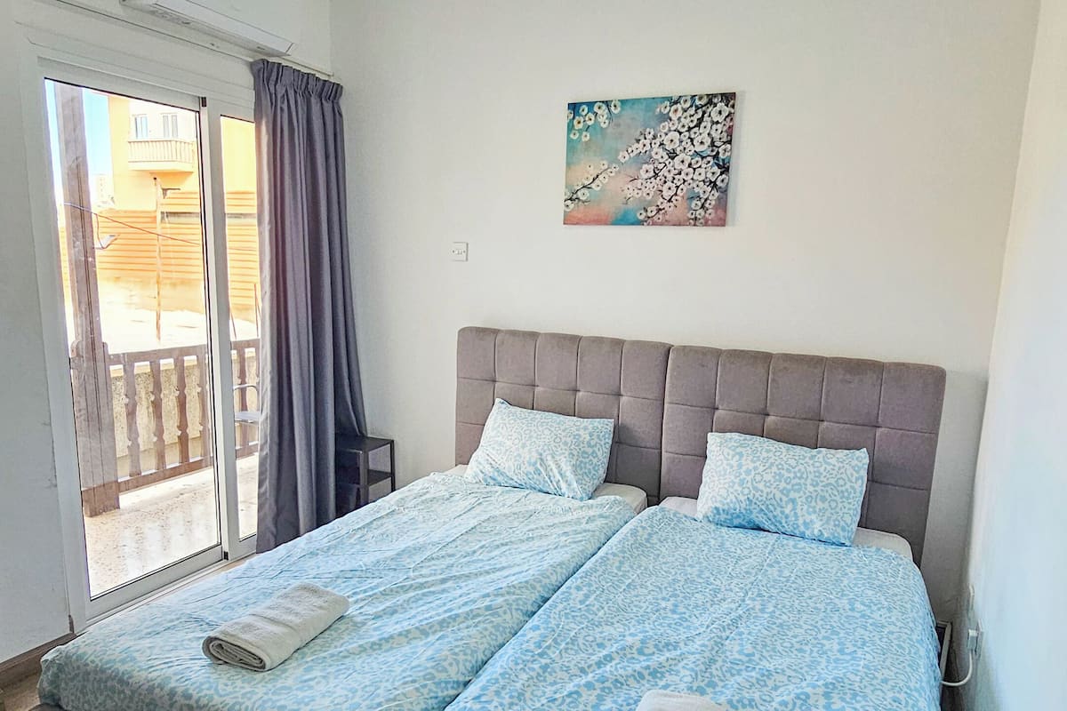 Uma cama de casal com cabeceira cinzenta e roupa de cama estampada em azul, um pequeno quadro na parede e uma janela com cortinas.