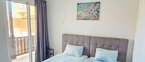 Un lit double avec une tête de lit grise et une parure de lit bleue à motifs, un petit tableau accroché au mur et une fenêtre avec des rideaux.