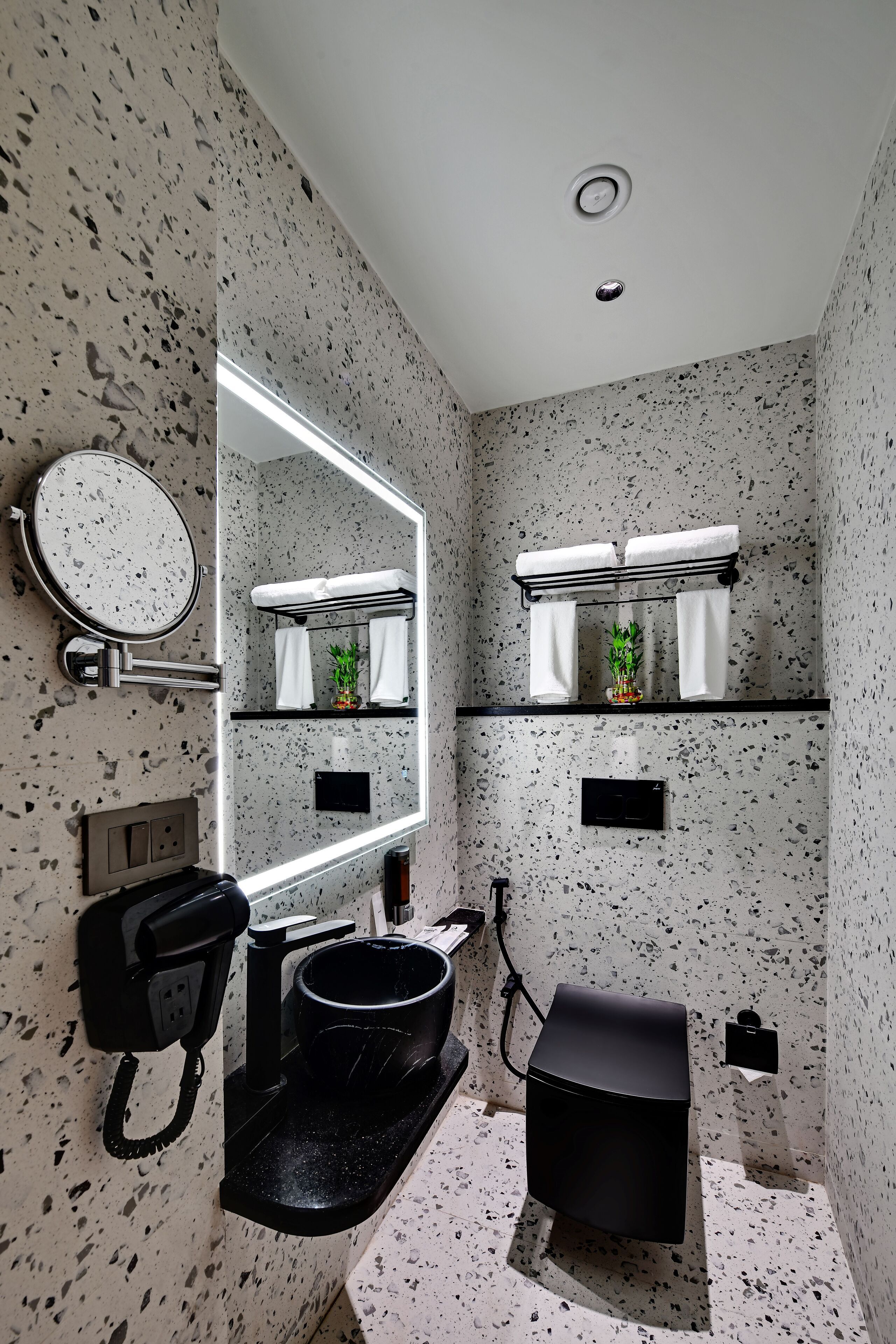 Un baño con salpicadero moteado, un espejo redondo, un lavabo negro, un mueble de tocador negro, un perchero con dos toallas y una pequeña planta en maceta.